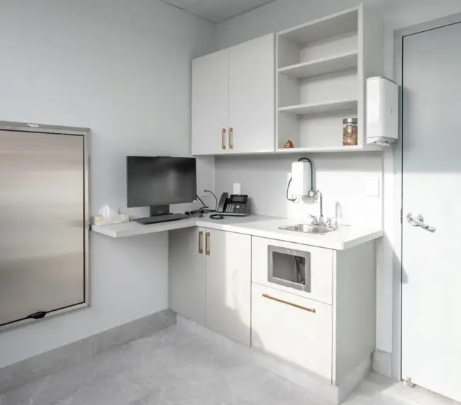 Small, sterile kitchenette/workstation in a pale gray room featuring a stainless steel fridge, white countertop with a computer monitor and phone, upper cabinets with open shelves, a sink, microwave drawer, and a door to the right.