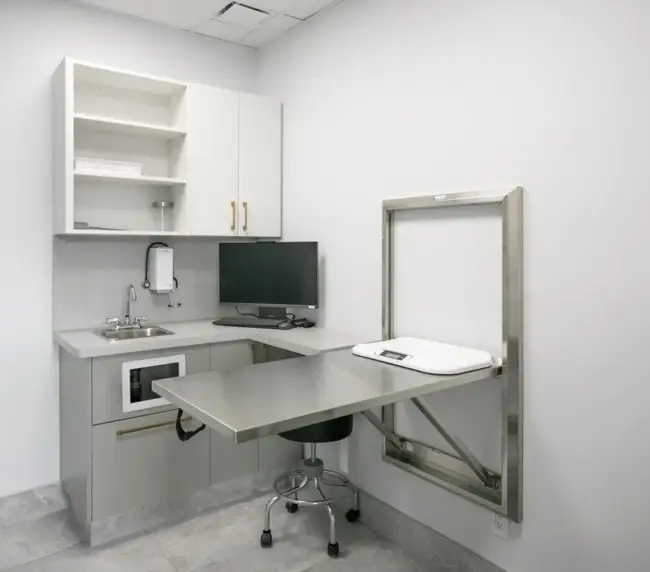 Radiology/diagnostic exam room with a stainless steel adjustable table, sink, computer monitor, and white wall cabinets.] ,