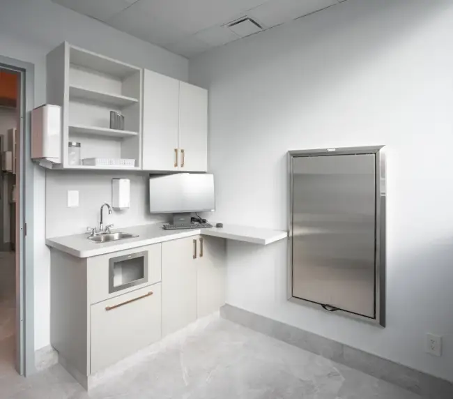 Compact kitchenette with white cabinets, a small sink, microwave inset, and open shelves beside a computer monitor.