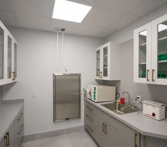Clinical kitchenette with white cabinets, a stainless steel pass-through, sink, microwave, and small sterilizing unit.