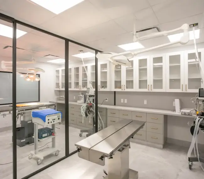 Modern hospital operating room with a metallic surgical table, overhead lights, glass partition, and anesthesia equipment.
