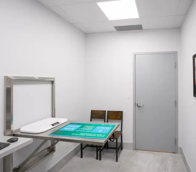 Medical imaging room with an X-ray table and a computer workstation, two wooden chairs, a door, and framed animal pictures on the wall