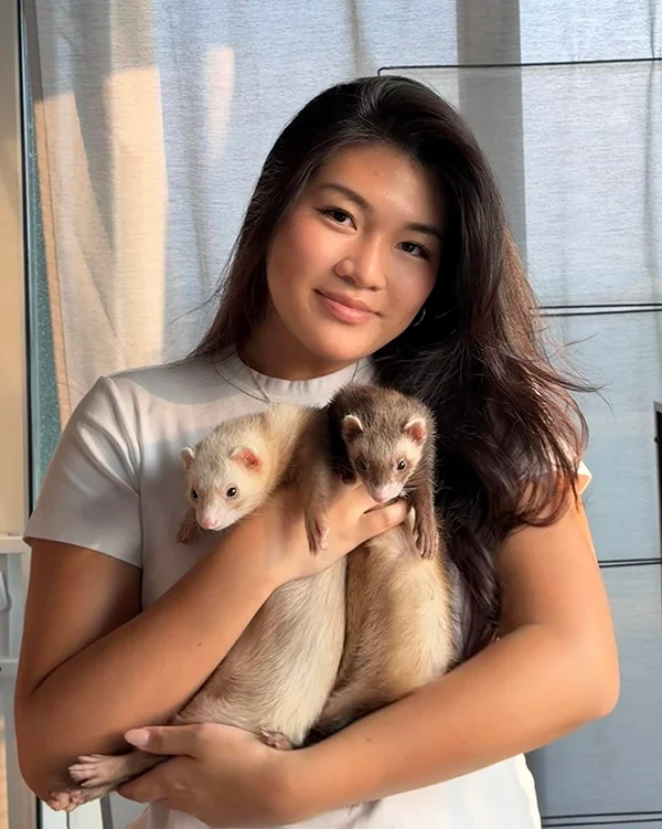 Woman smiles softly while holding two ferrets, one cream-colored and one brown, against a light indoor backdrop near a window.