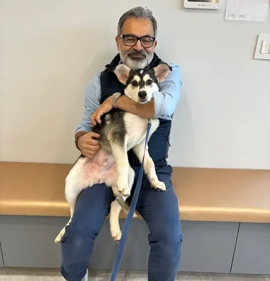 Man with glasses sits on a clinic bench, hugging a small black-and-white dog on his lap.