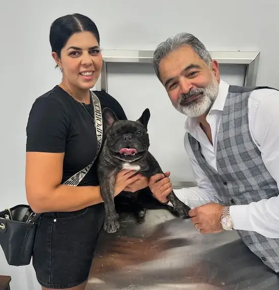 Two people pose with a small black French bulldog on a metal exam table, smiling at the camera in a clinic setting.